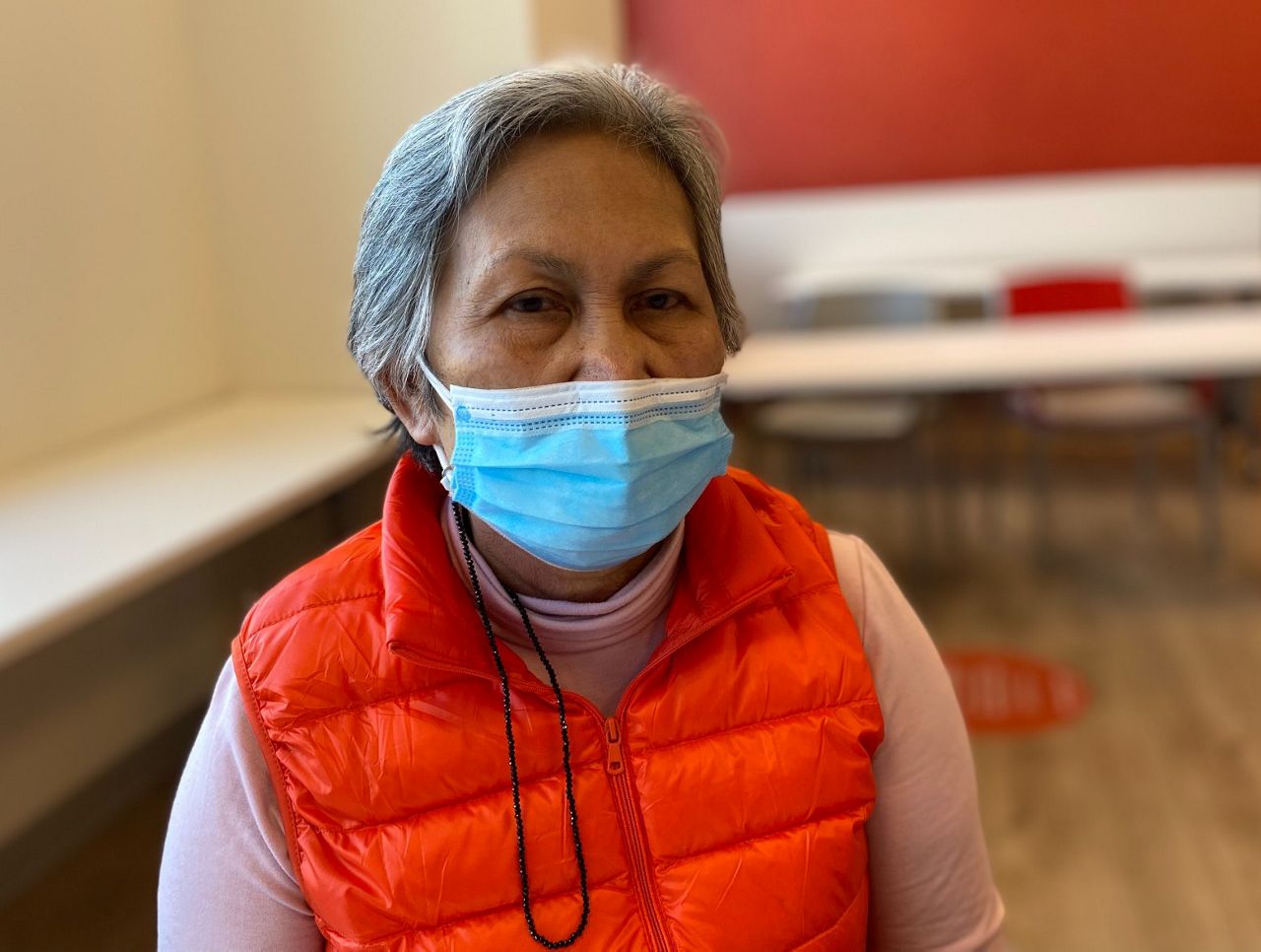 Portrait-style photo of Elvie Roman, sitting in a red puffer vest and a blue surgical mask on.