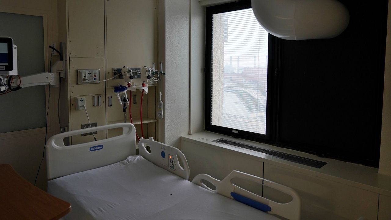 An empty hospital room with a hospital bed in center and machinery attached to the wall, window blinds halfway open on the other wall