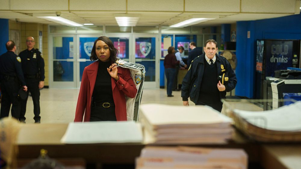 The actress Amanda Warren and the actor Richard Kind walk towards the viewer inside a police precinct station, with a stack of papers on a desk in the foreground.