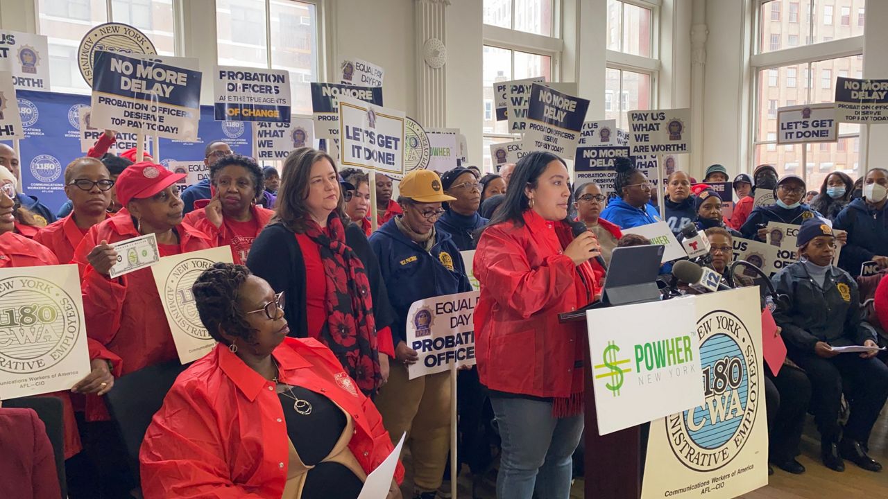 Crowd of advocates holding equal pay-related signs behind a row of seated elected officials, with Amanda Farias standing and speaking at the podium in the center 