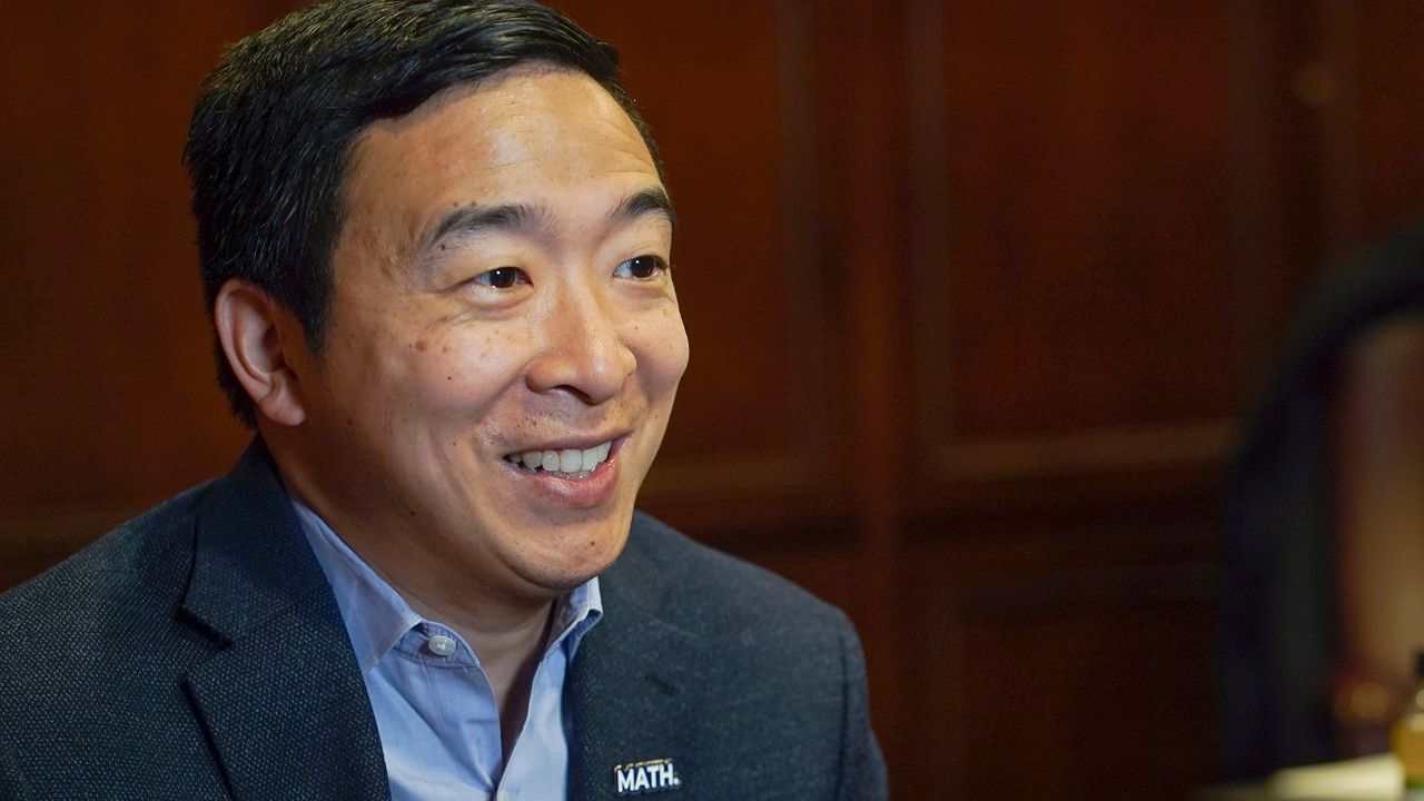 Andrew Yang, wearing a black suit jacket and a sky blue dress shirt, sits in a room.