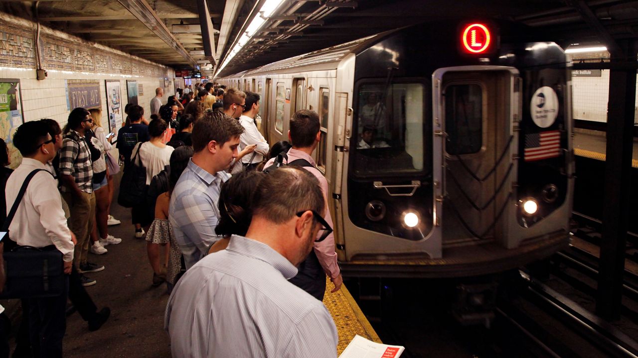A silver train, right, with a black cover, headlights, and a red "L" inside a red circle on its temple, is about two feet away from a yellow platform. A crowd of people stand on the platform.