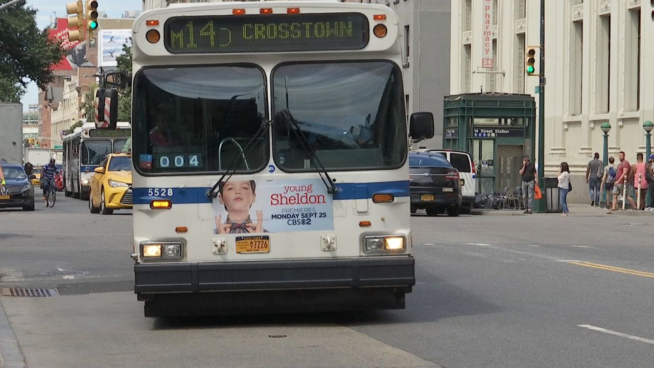 A white bus with a black bumper driving on a black road. Green text on the temple of the bus reads "M14 Crosstown"