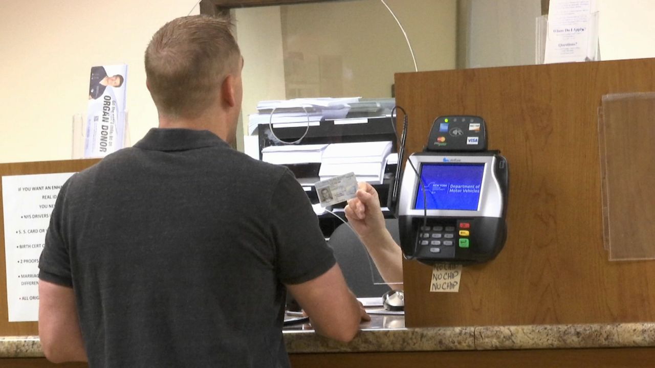 A person, wearing a black-collared shirt, stands at a brown marble counter, near a debit card machine. A person holds up a white driver's license at at clear screen.