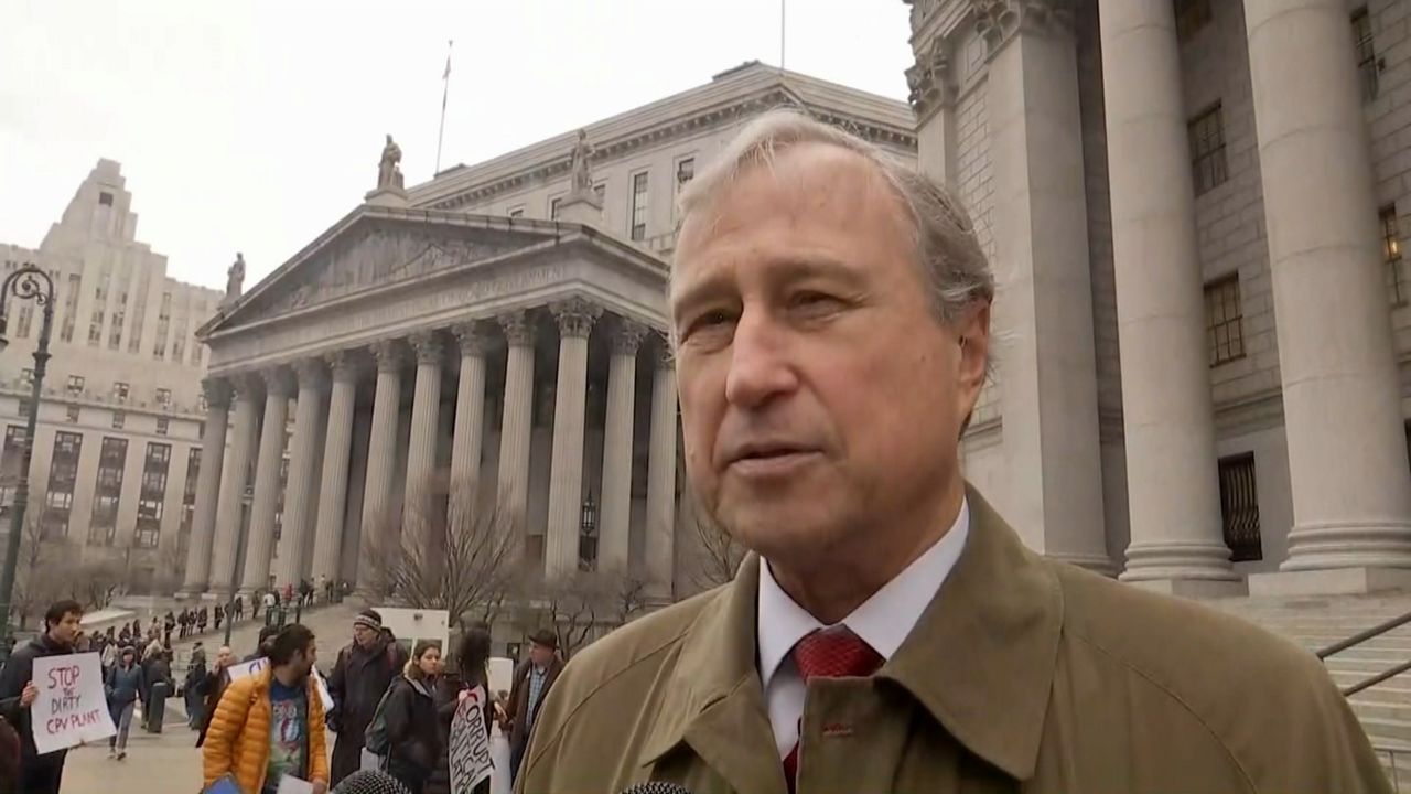 A man, wearing a brown coat, a white dress shirt, and a red tie, stand in front of a crowd and several white marble courthouses.