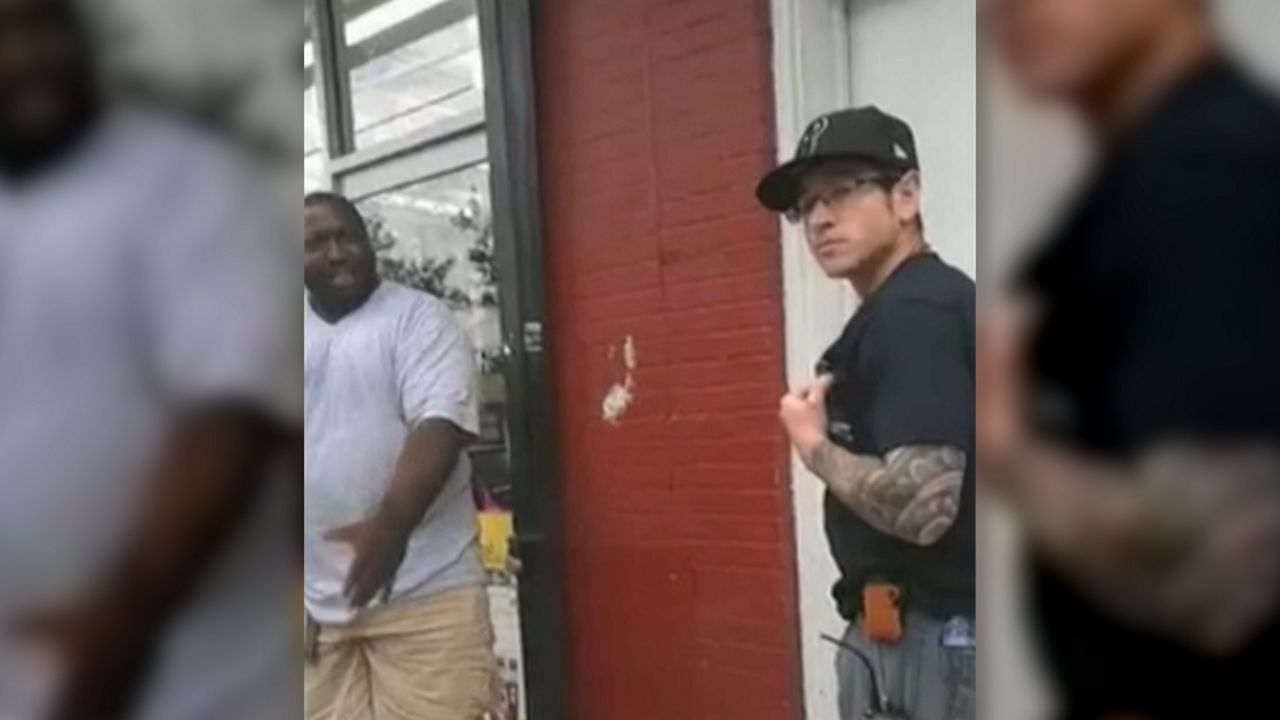 A man, right, wearing black square glasses, a black Yankees baseball cap, a black tshirt, and blue jean pants, holds his shirt while standing about 10 feet to the left of Eric Garner, wearing a white tshirt and grey khakis. A red door stands between them.