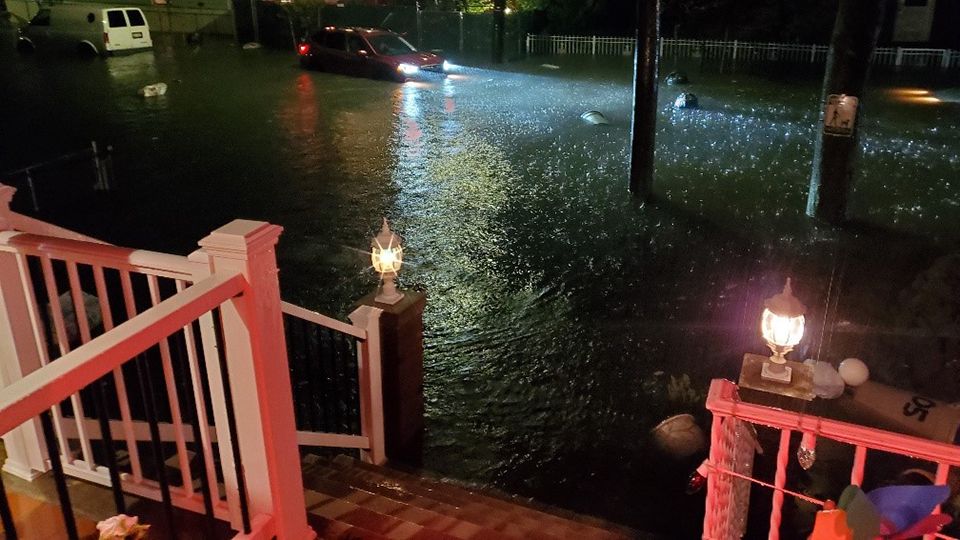 Looking down from a white wooden porch in Queens at night, the street is completely flooded with water, as a car tries to drive through it. 