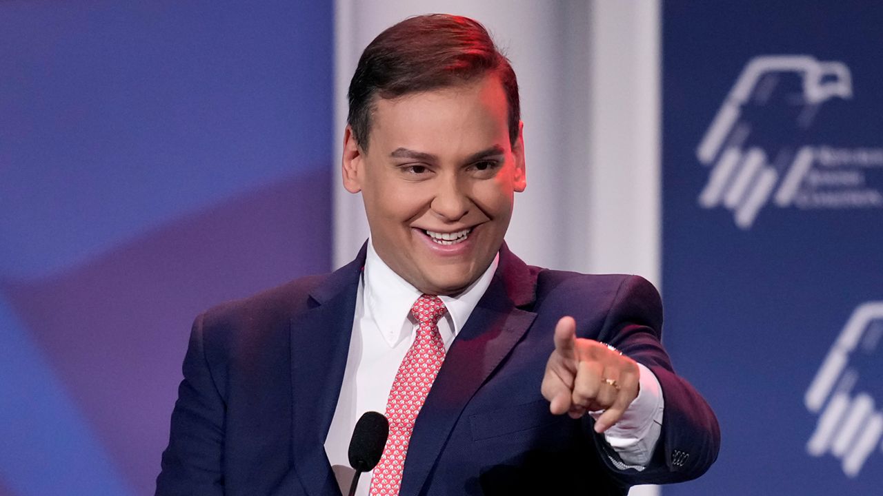 George Santos, wearing a navy suit, white collared shirt and a bright pink tie, points at the camera while speaking at a podium in front of a wall bearing the logo for the Republican Jewish Coalition.