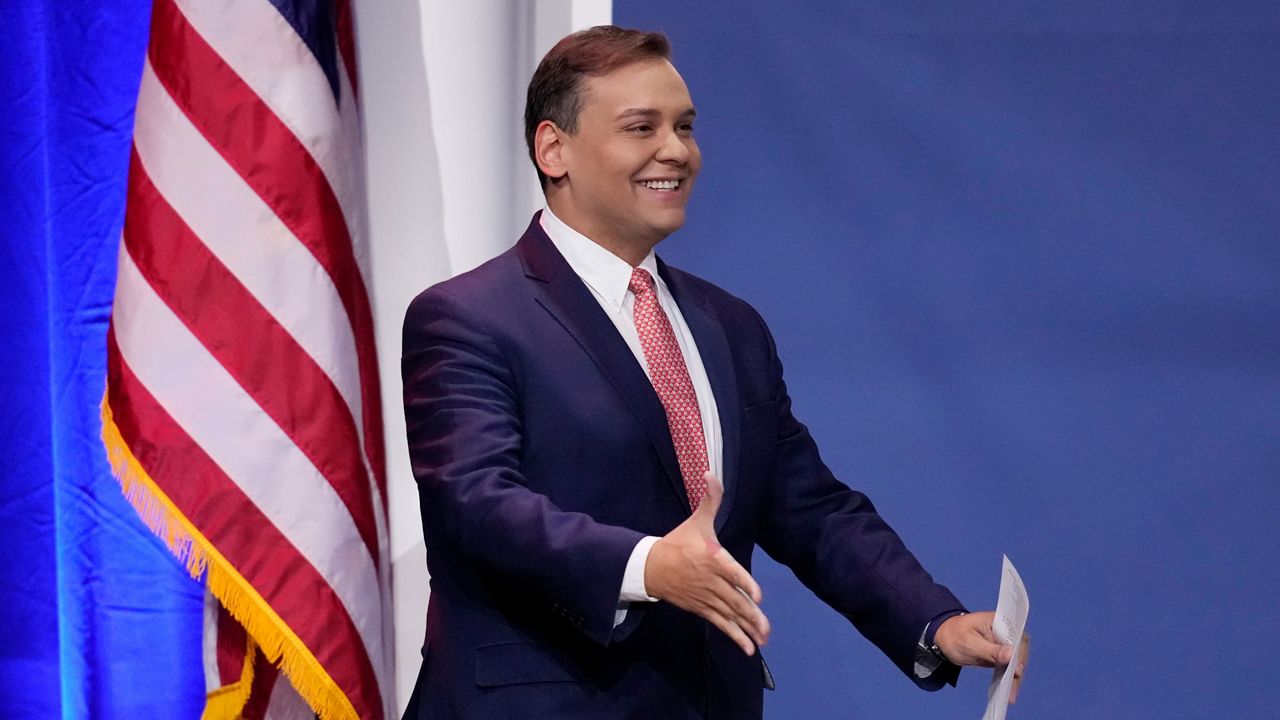 Rep.-elect George Santos is pictured in a navy suit, white shirt and pink tie in front of a blue background and an American flag.