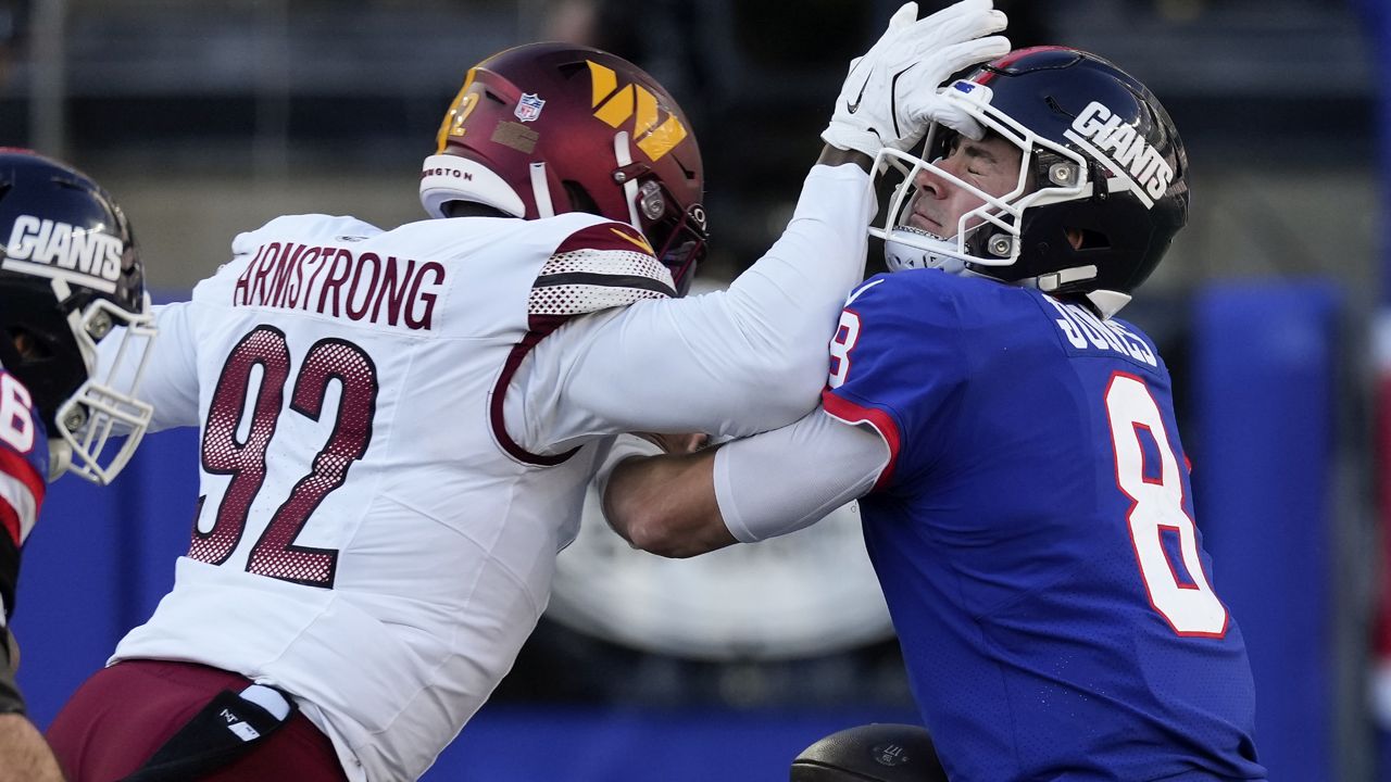 washington commanders defender, in white jersey, hits new york giants quarterback, in blue throwback jersey, with forearm before sacking him