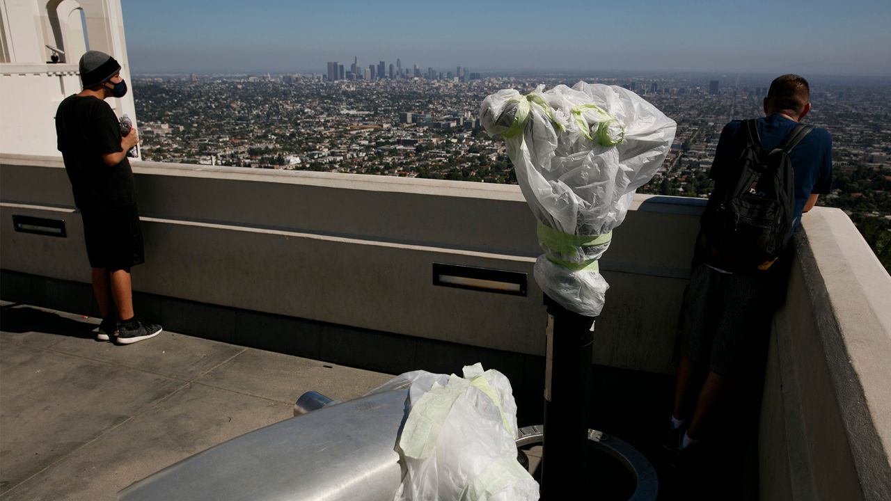 Two people stand near telescopes wrapped in plastic to prevent the spread of COVID-19 at the Griffith Observatory overlooking downtown Los Angeles, Wednesday, July 15, 2020. Coronavirus cases have surged to record levels in the Los Angeles area, putting the nation's largest county in "an alarming and dangerous phase" that if not reversed could overwhelm intensive care units and usher in more sweeping closures, health officials said Wednesday. (AP Photo/Jae C. Hong)