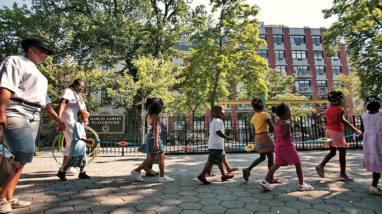 A line of children are seen walking down the sidewalk with a caretaker behind them and a playground in the background.