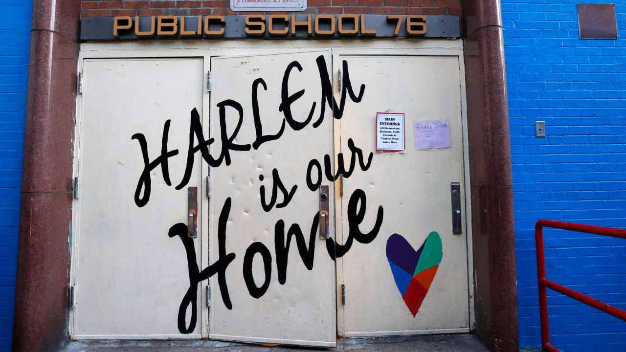 White double doors to a school has the words "Harlem is our home" written on it with a multicolored heart on bottom right. On top of doors there is a sign with name of school "Public School 76" 