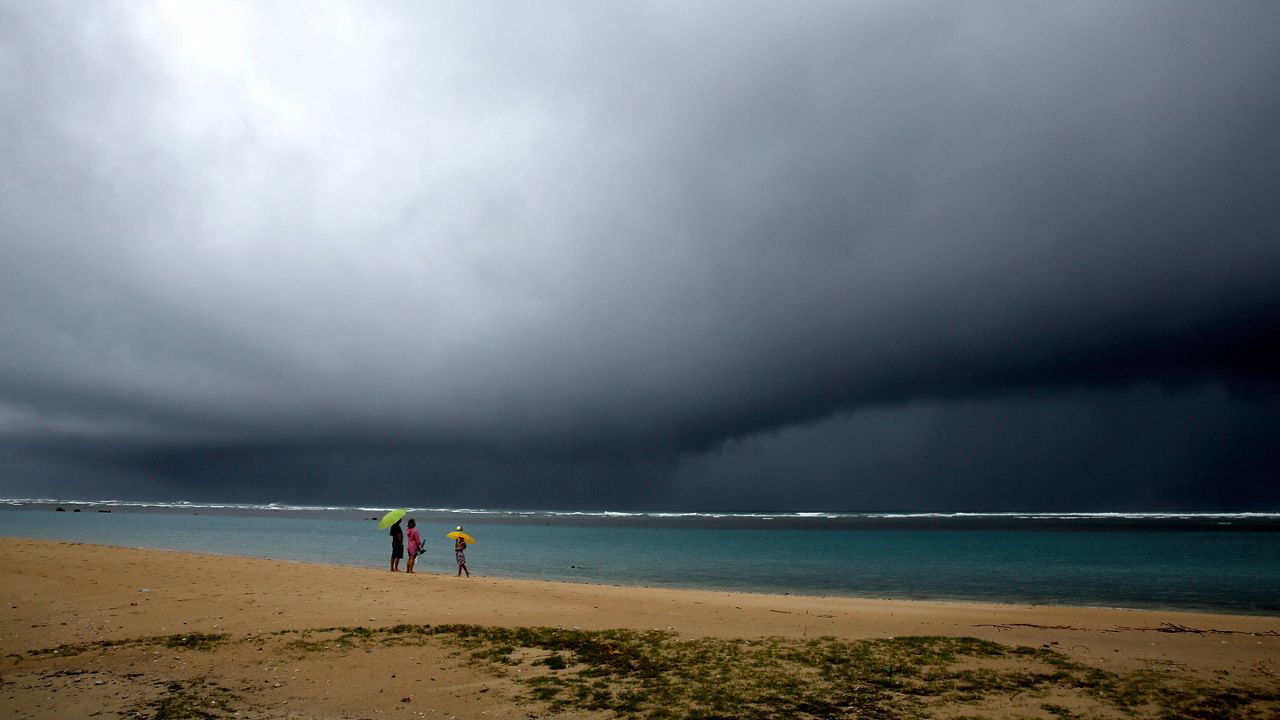 People hold umbrellas as it begins to rain on an otherwise empty beach in Honolulu on Monday, Dec. 6, 2021. A strong storm packing high winds and extremely heavy rain flooded roads and downed power lines and tree branches across Hawaii, with officials warning Monday of potentially worse conditions ahead. (AP Photo/Caleb Jones)