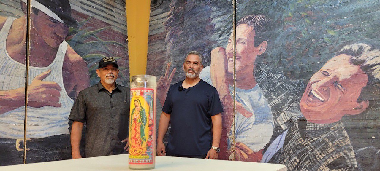 (Left to right) Bert Hernandez and Armando Hernandez stand in front of their late brother Adan Hernandez’ mural from the Chicano film “Blood In, Blood Out.” (Photo credit: Jose Arredondo)