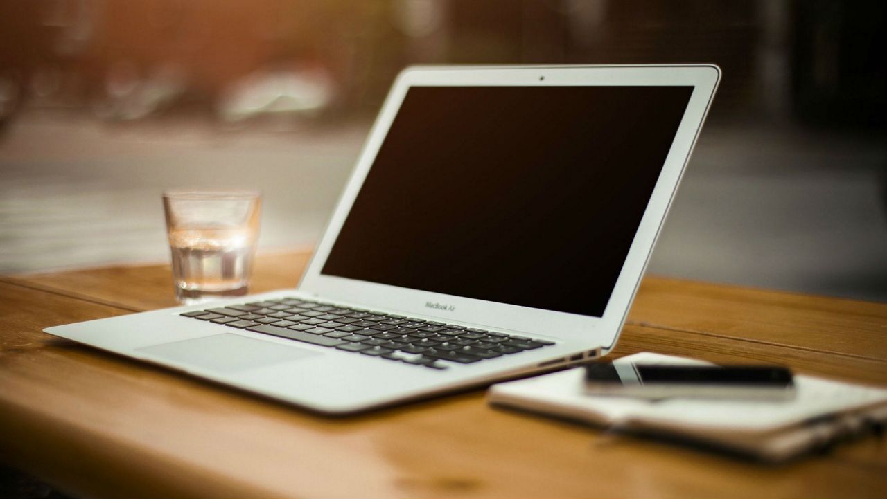 A white laptop rests on a brown wooden desk, next to a white notebook on which a black pen rests. A clear glass sits on the table, to the right of the laptop.