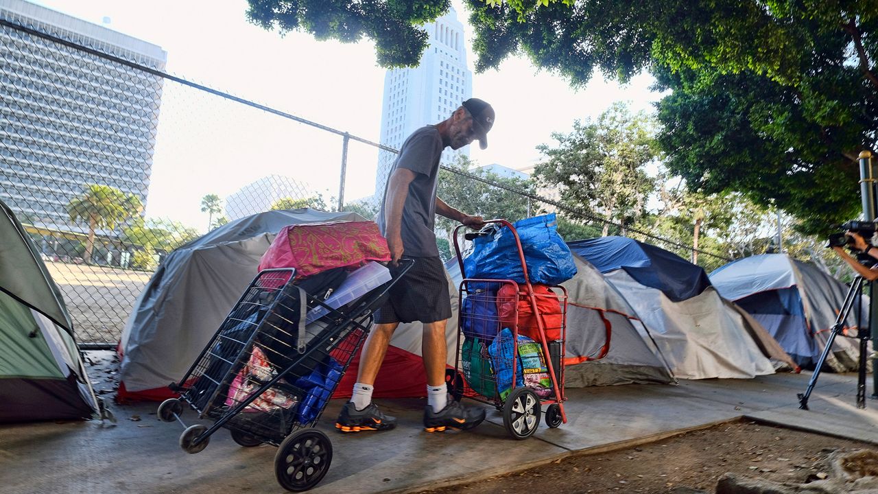 In this July 1, 2019 file photo, a homeless man moves his belongings from a street near Los Angeles City Hall, background, as crews prepared to clean the area. (AP Photo/Richard Vogel, File)