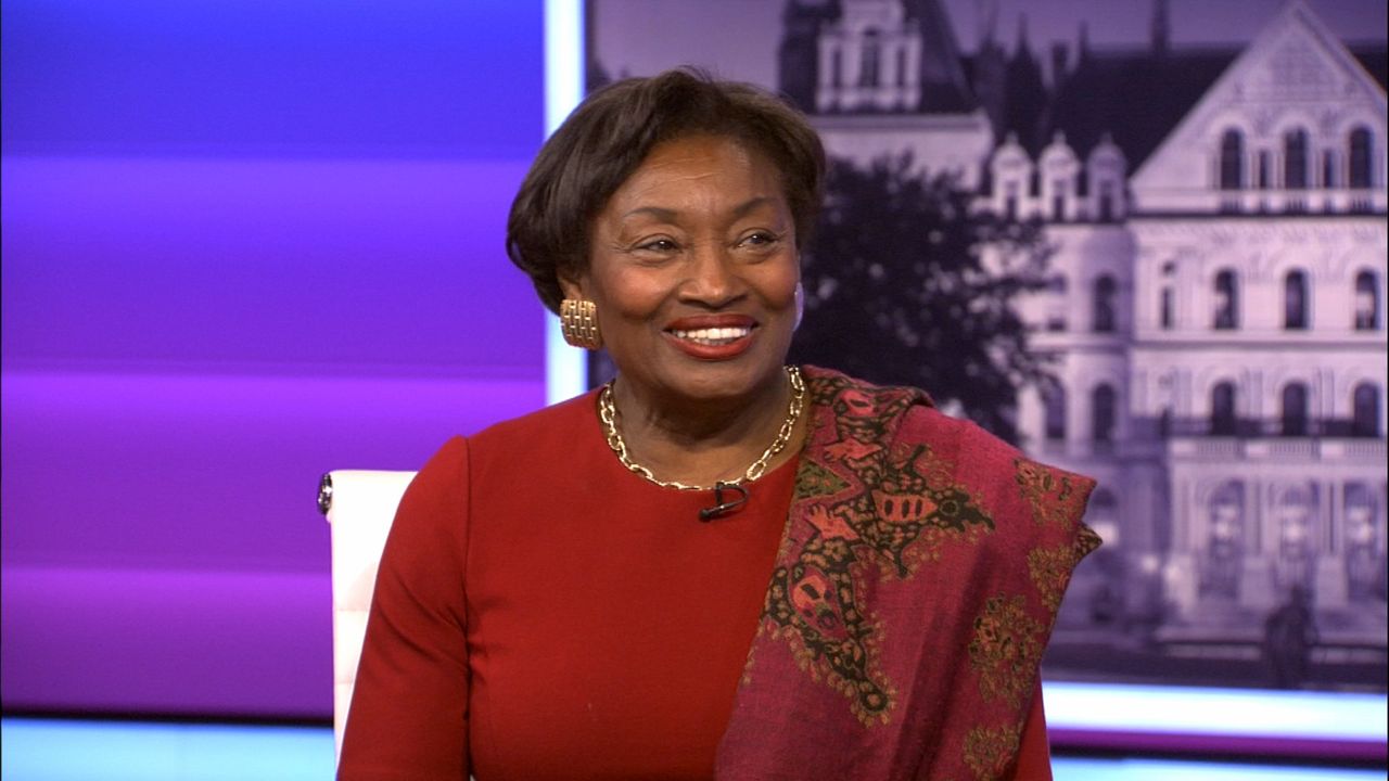 Andrea Stewart-Cousins, wearing a red blouse and a red shawl, sits in a white chair against a purple background on the set of NY1 political show "Inside City Hall."
