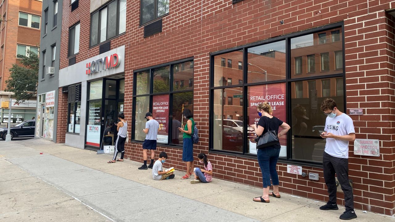 Five people standing socially-distant, looking at their phones, outside a CityMD clinic.