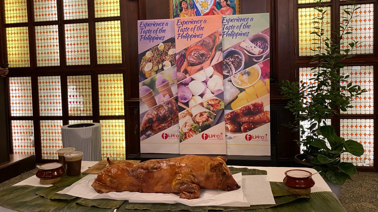 A plate of lechon on top of bamboo leaves atop a table with banners behind that read "Experience a Taste of the Philippines"