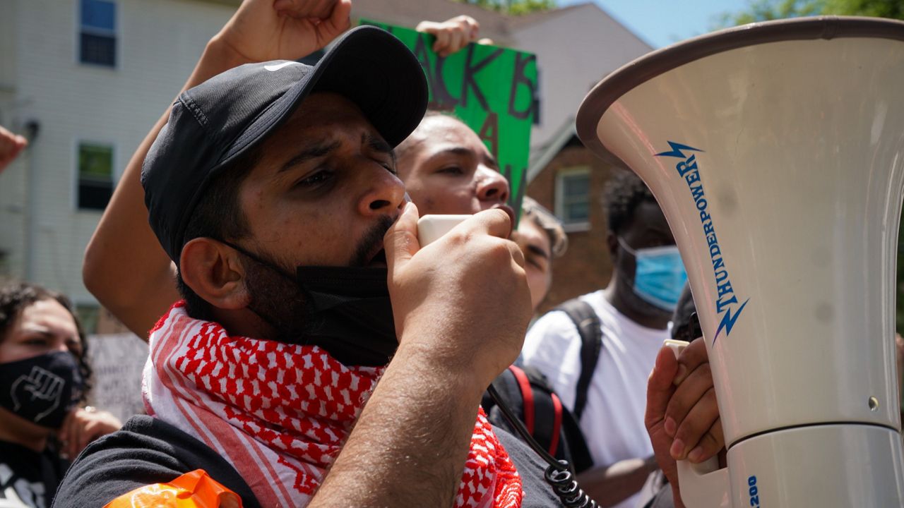 Mohammad wearing a black shirt, red scarf around his neck, and a black cap with a megaphone in hand with other protesters behind him.