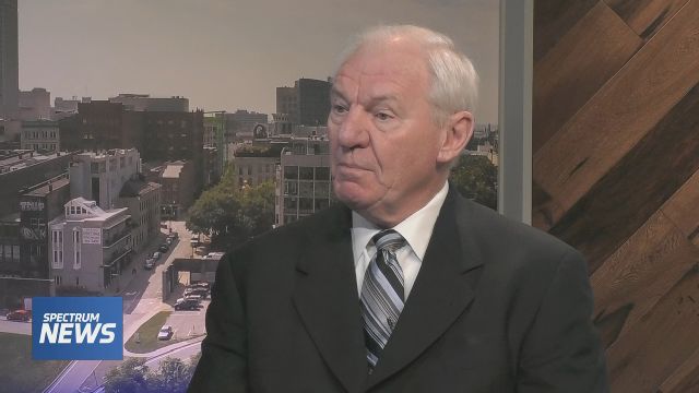 A man wearing a black suit jacket, a white dress shirt, and a multi-colored tie sits against wallpaper depicting a city on the set of 