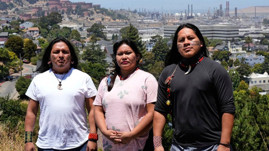 Juan Bay, Jhajayra Machoa Mendúa y Nadino Calapucha, indígenas de Ecuador posan mientras se ve una refinería de Chevron al fondo.