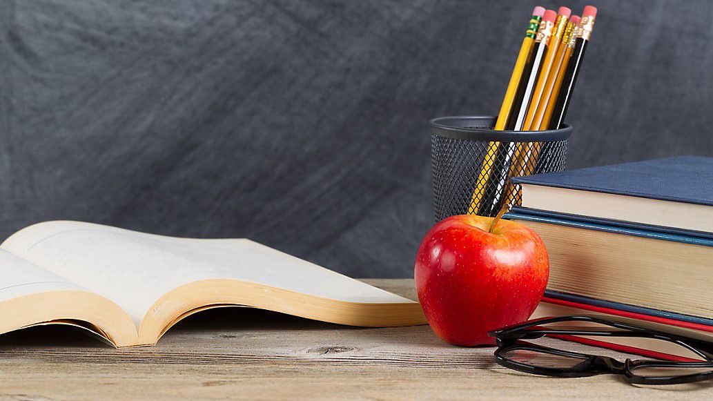 Desktop with books, red apple, reading glasses, and pencils in front of blackboard. Layout in horizontal format with plenty of copy space. (iStock image)