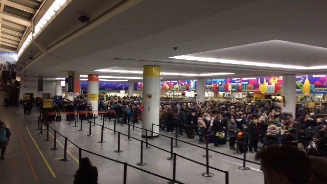 A large crowd of people stands close together near concrete pillars. Everyone has luggage and is dressed warmly.