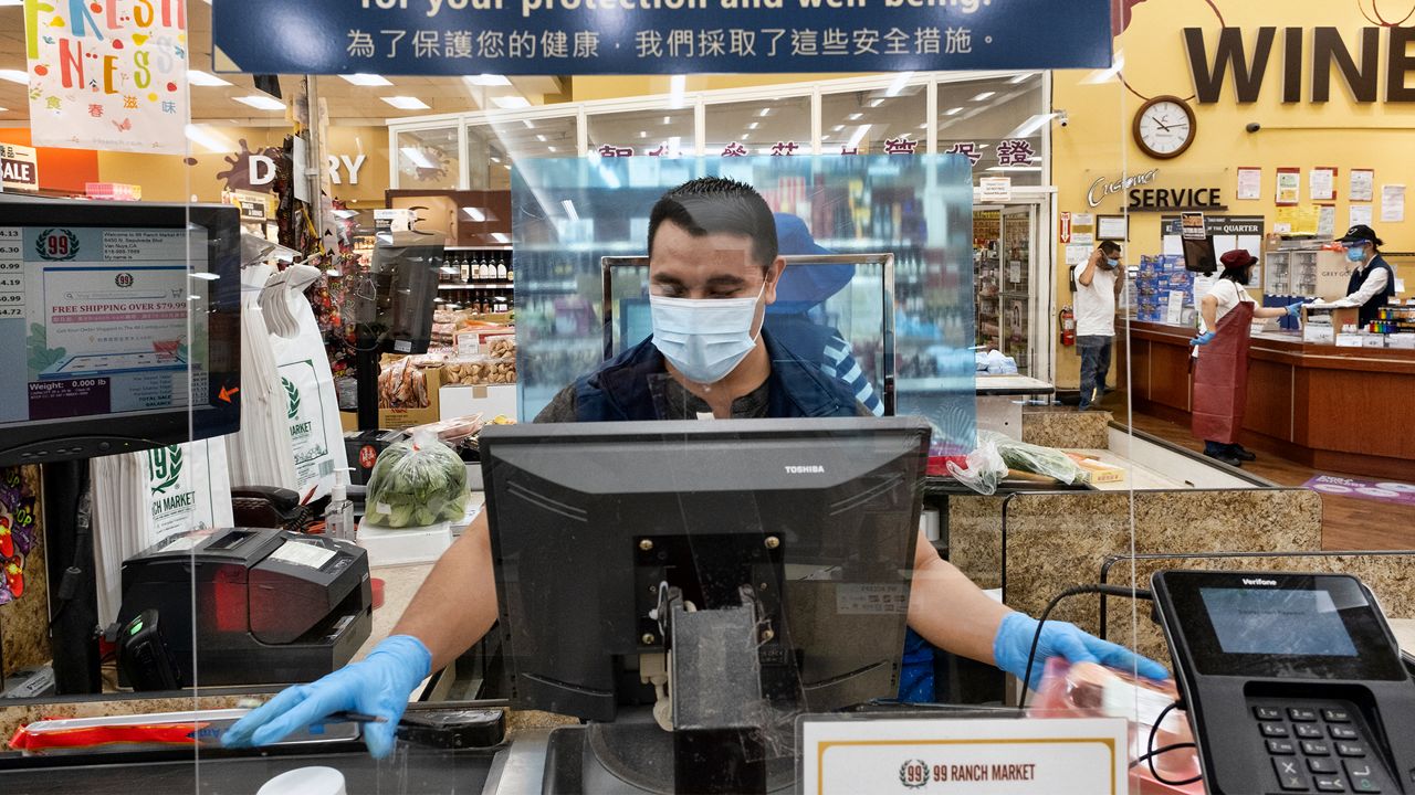 A grocery worker, wearing a protective mask and gloves, as he helps to check out a customer from behind a plexiglass barrier at the 99 Ranch Market in the Van Nuys section of Los Angeles on Tuesday May, 5, 2020. (AP Photo/Richard Vogel)