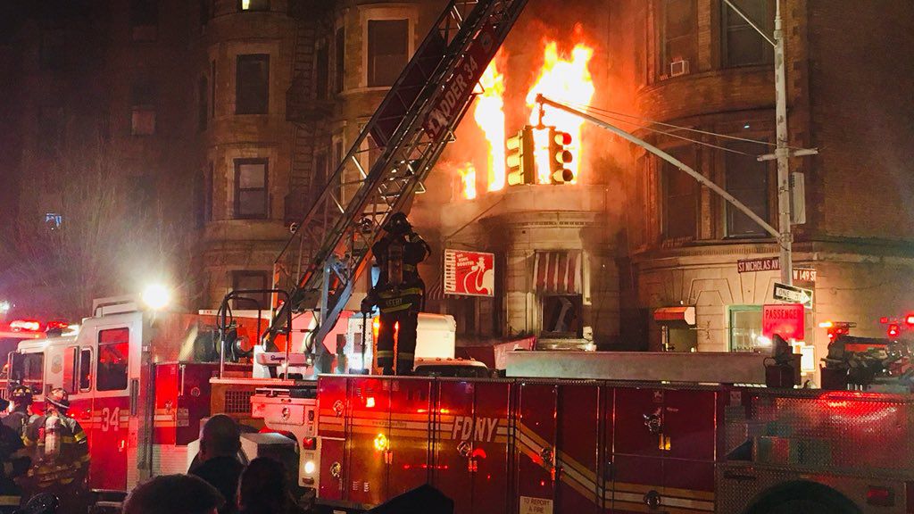 A firetruck, with a ladder and firefighters, outside a building. Orange flames are spewing out of one of the building's windows.