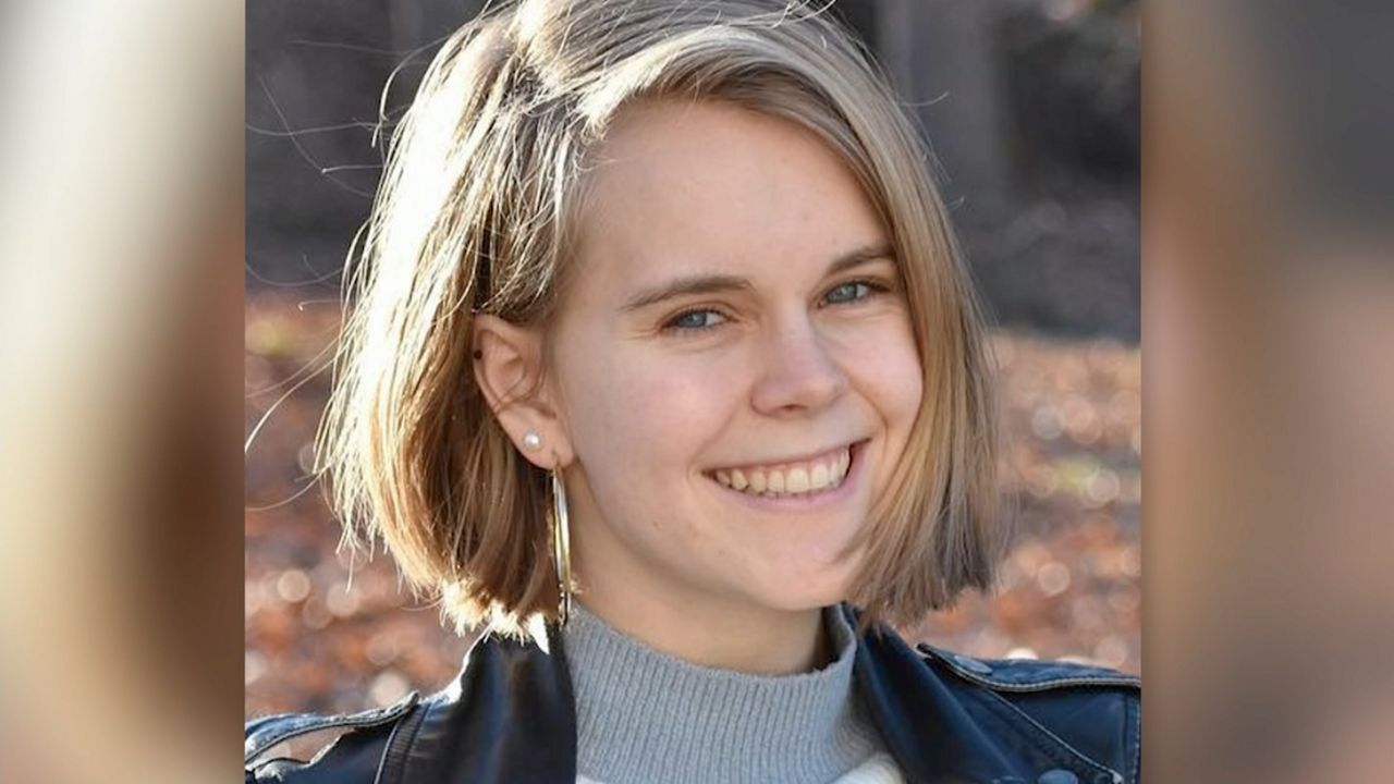 A woman, wearing a blue jean jacket, a grey sweater, and earrings.