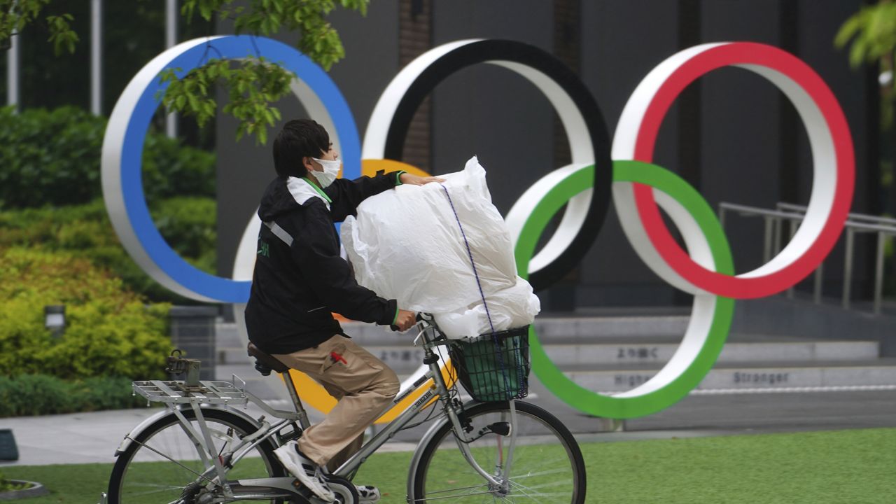 FILE: An employee wearing protective masks to help curb the spread of the coronavirus rides a bicycle in front of he Olympic Rings Thursday, May 13, 2021, in Tokyo. (AP Photo/Eugene Hoshiko)