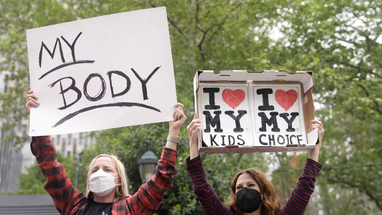 Two protesters hold signs that read "My Body" and "I 'heart' my kids, I 'heart' my choice" with trees seen behind them.