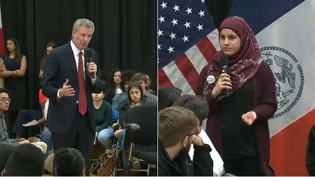 New York City Mayor Bill de Blasio, right, wearing a black suit, a white dress shirt, and a red tie while holding a black microphone in his left hand; a girl wearing a red hijab, a black shirt, and a red fleece while holding a black microphone in her right hand.