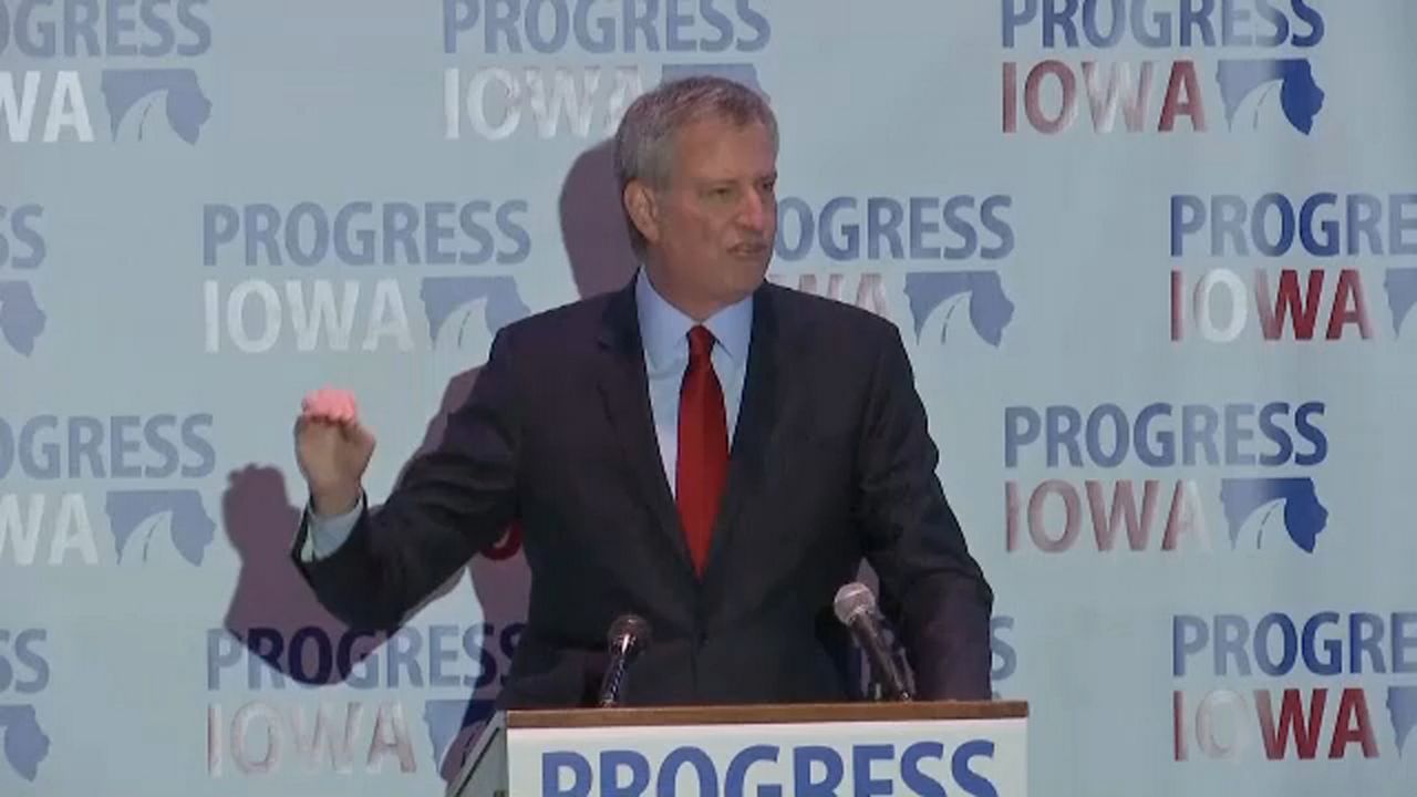 New York City Mayor Bill de Blasio in a black suit, with a red tie and a blue shirt. He stands at a podium with two microphones. Signs for "Progress Iowa" litter wallpaper behind him.