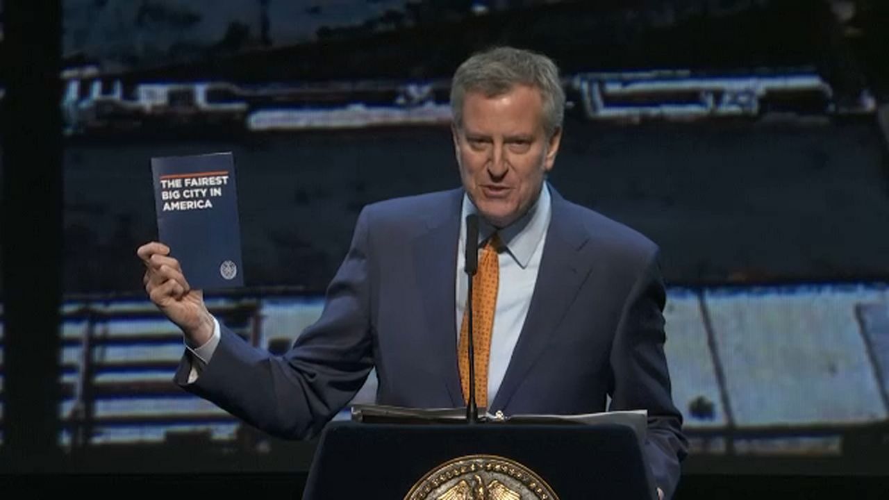 New York City Mayor Bill de Blasio, wearing a dark blue suit, a white dress shirt, and an orange-spotted tie, stands into a black microphone on a blue lectern. He holds a blue book with text on its cover that reads "The fairest big city in America."