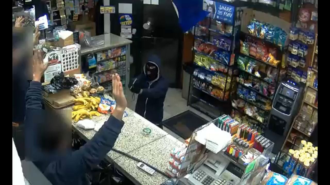 Two people behind a marble counter put their hands in the air as a person wearing a blue hoodie and a partial mask points something at them. Groceries are in the background, and a cash register is to the right of one of the people who has their hands in the air.