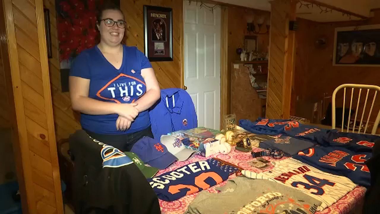 A girl wearing glasses a blue shirt stands against a brown wall. A variety of pinstriped, blue, and grey Mets jerseys and shirts are folded and rest on a table. A white chair is at the end of the table.