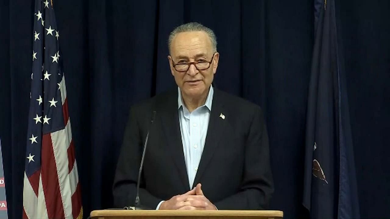 Charles Schumer wearing a black suit, a white dress shirt, and rectangular glasses. His hands are folded, resting on a beige lectern. A black microphone is a few inches to the right of his hands. An American flag is about two feet to his right. Dark blue curtains are behind him.