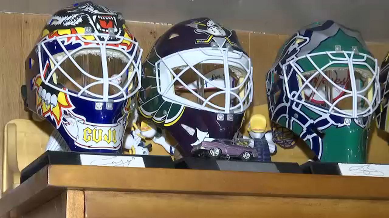 Three hockey goaltender masks rest on black plaques on top of a brown cabinet. On the left is a yellow and black mask, in the middle is a black Mighty Ducks mask, and on the right is a green mask.