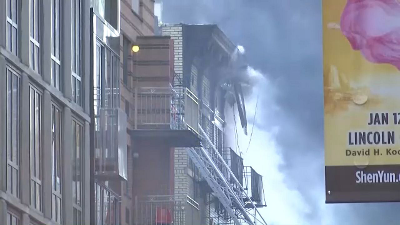Black and grey smoke billows from the top of a dark building. The building is three-windows-wide. A white fire department ladder rests on the front of the building.