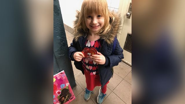 A girl in a red dress, adorned with the design of hearts, wears a blue winter coat and aqua sneakers.