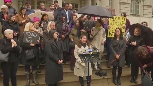 A group of people dressed in coats, holding umbrellas and signs, on the steps of a building. A small podium with three microphones stands before a woman in a white raincoat.