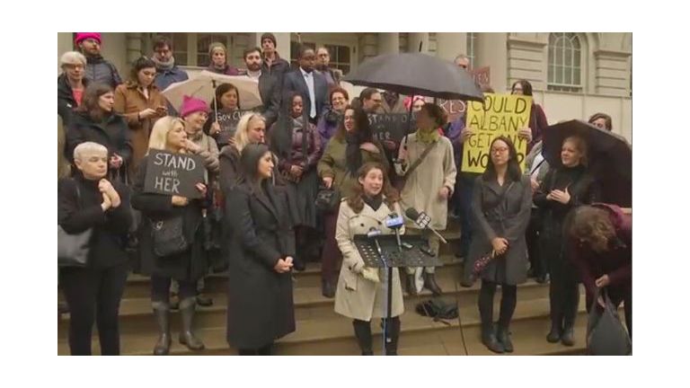 A group of people dressed in coats, holding umbrellas and signs, on the steps of a building. A small podium with three microphones stands before a woman in a white raincoat.