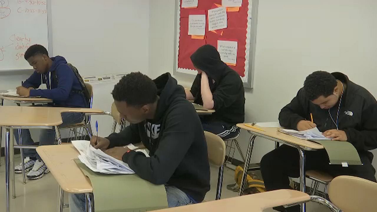 Four teenagers sit on single-seat brown desks in a classroom. A white wall is behind them. They have exam sheets on their desks.