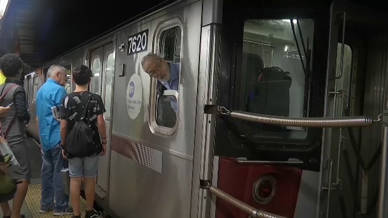 A man, center, wearing glasses and a blue shirt pokes his head out of a window on a silver subway car. Three people stand about three feet away from the man in front of doors to a subway car.