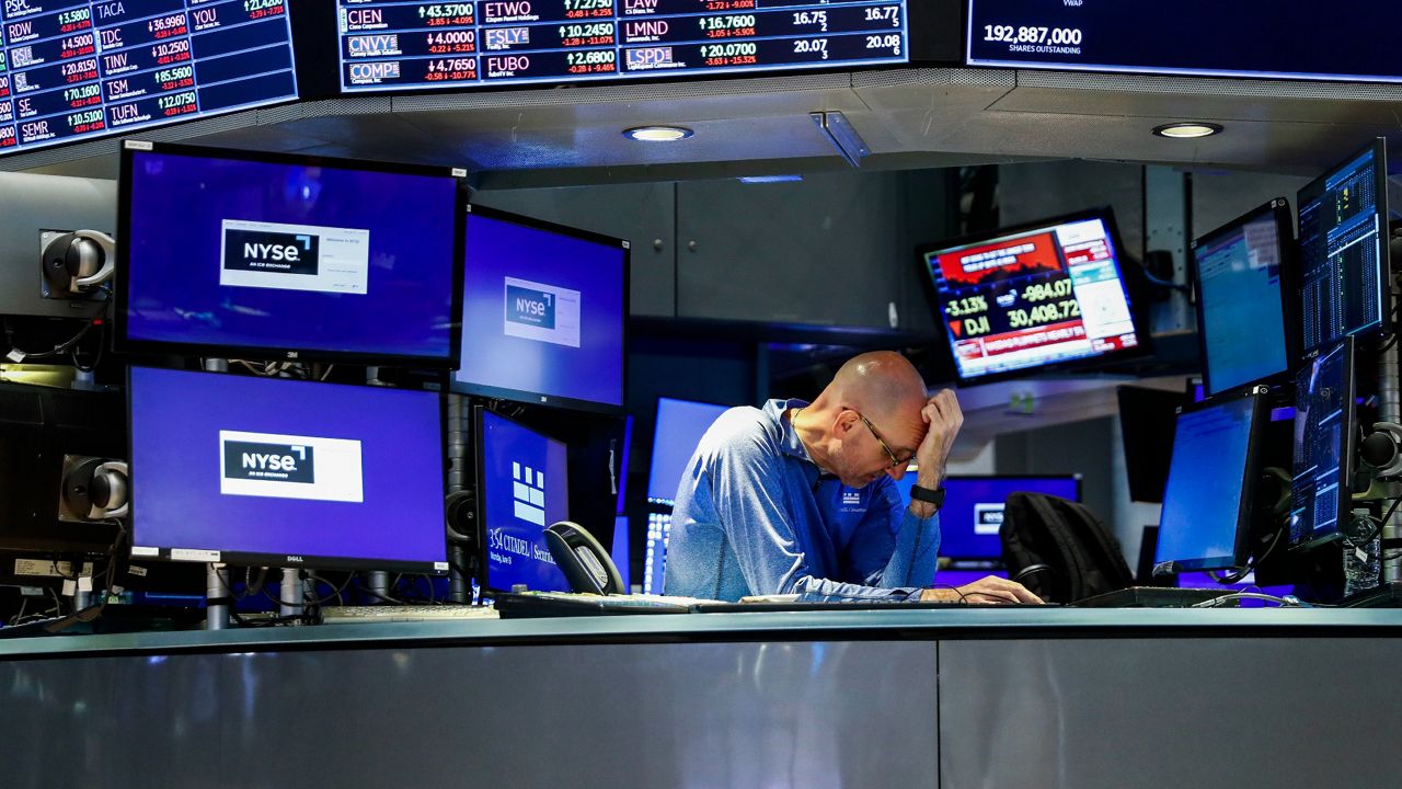 A trader holds his head in his hand, looking distressed, at a desk on the floor of the New York Stock Exchange with computer monitors all around him. 