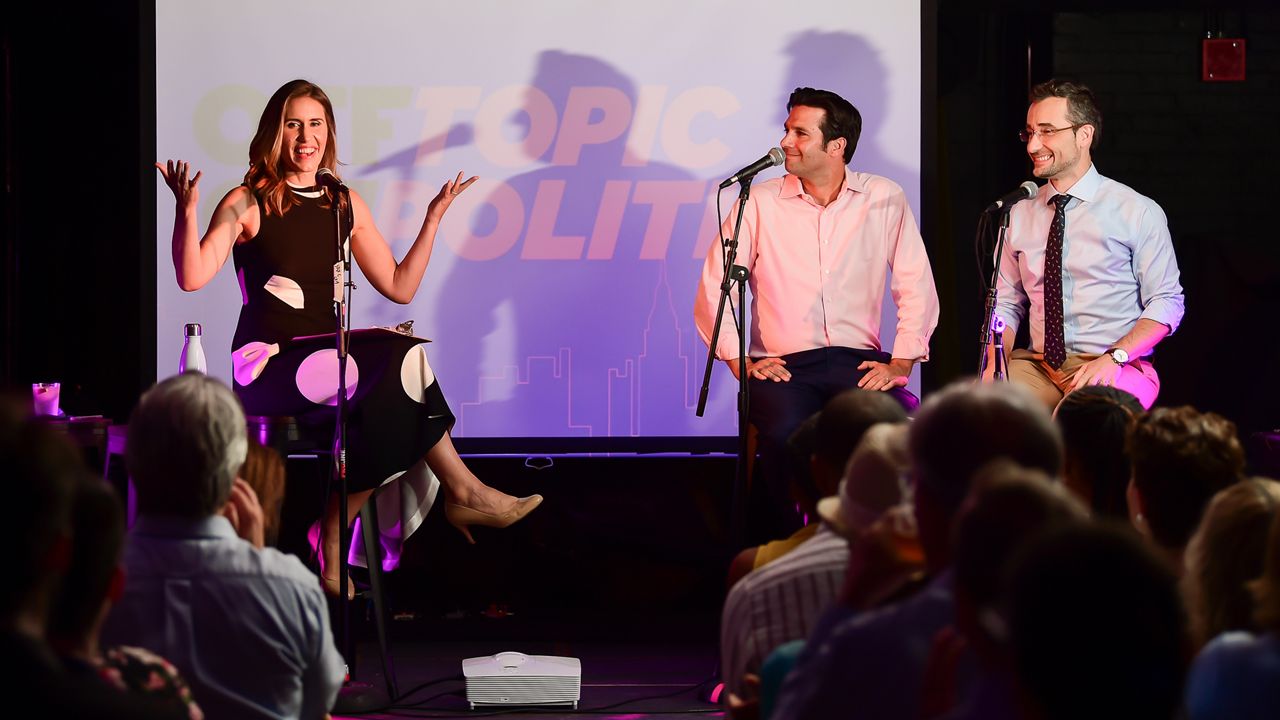 From left to right: Grace Rauh, Zack Fink, and Juan Manuel Benítez sit on a stage with black microphones in front of them. The "Off Topic/On Politics" logo, with black and white text in bold, is emblazoned on a purple projection on a white screen against a dark background. A crowd of people sit in the foreground.