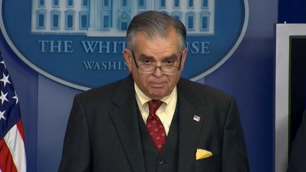 A man wearing a black suit, a black vest, a yellow shirt, a scarlet tie, and rectangular-shaped glasses strands in front of a blue wallpaper, an American flag, and a White House logo.
